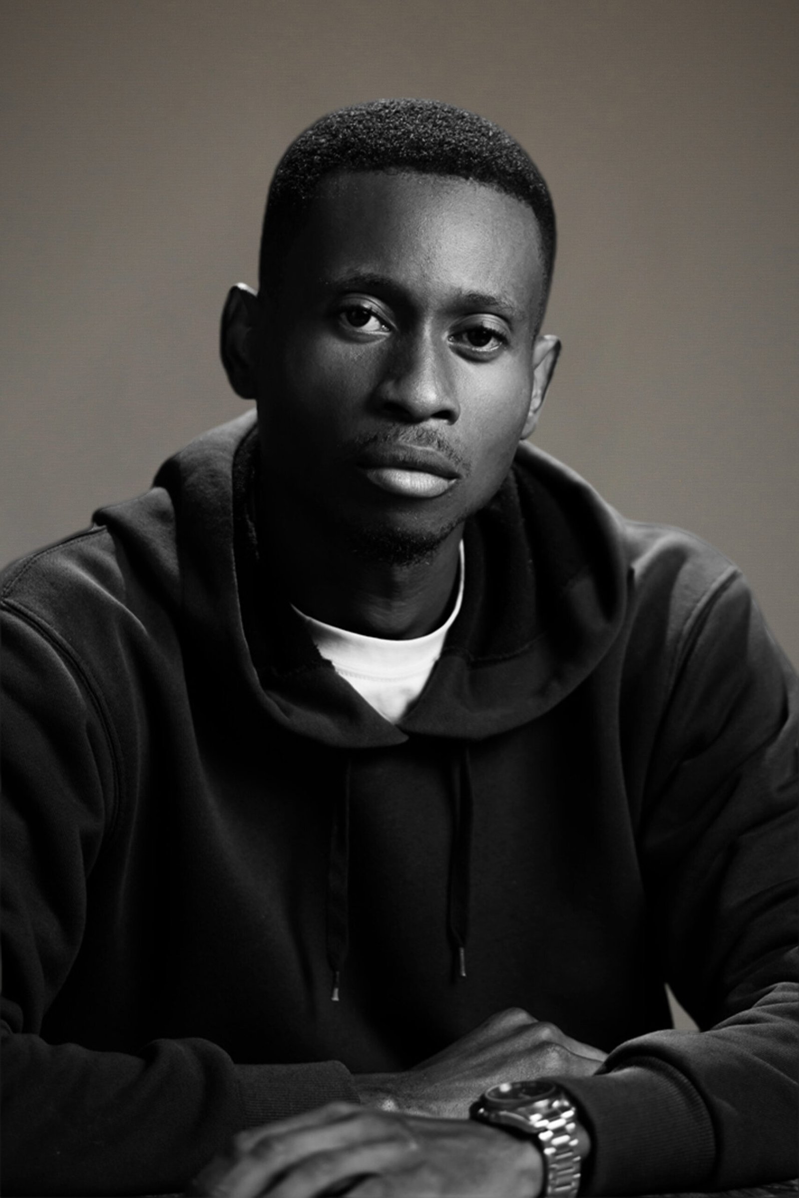 A man in a dark hoodie and watch sits with his arms resting in front of him, looking directly at the camera with a neutral expression against a plain background.