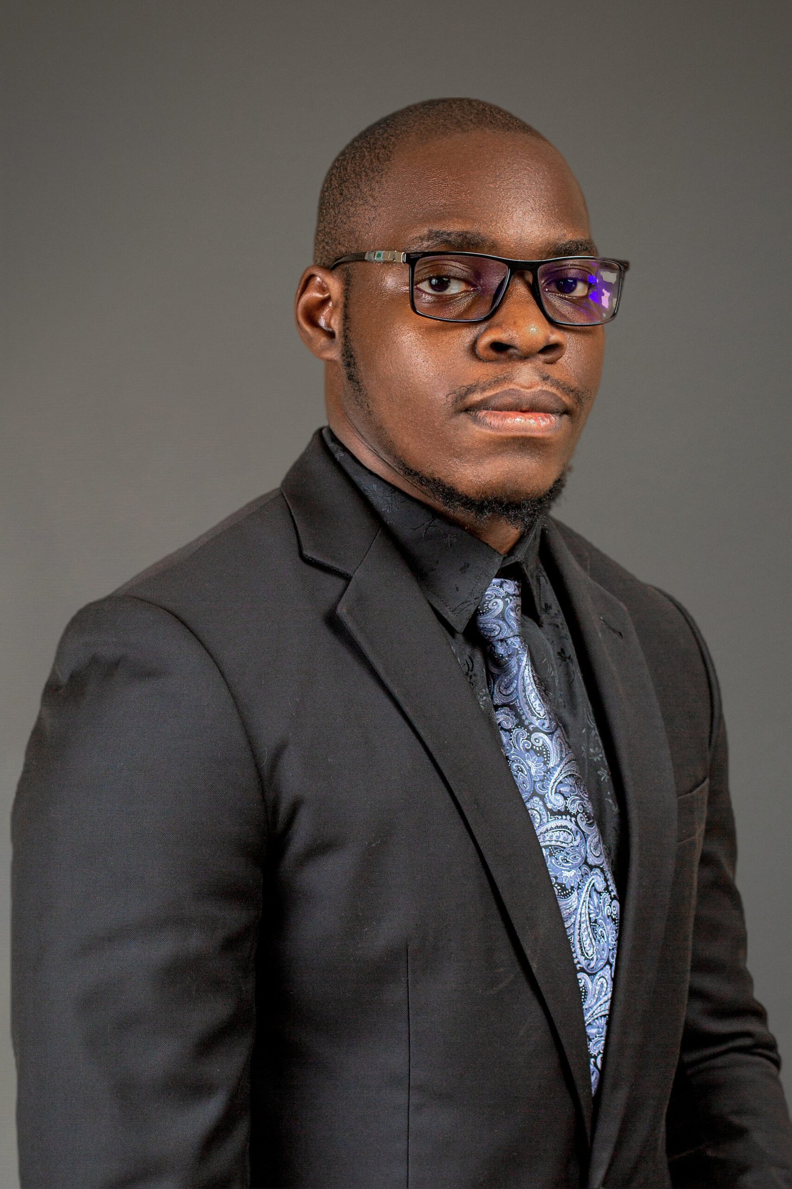 A man in a black suit, patterned tie, and glasses poses in front of a plain gray background, looking confidently at the camera.