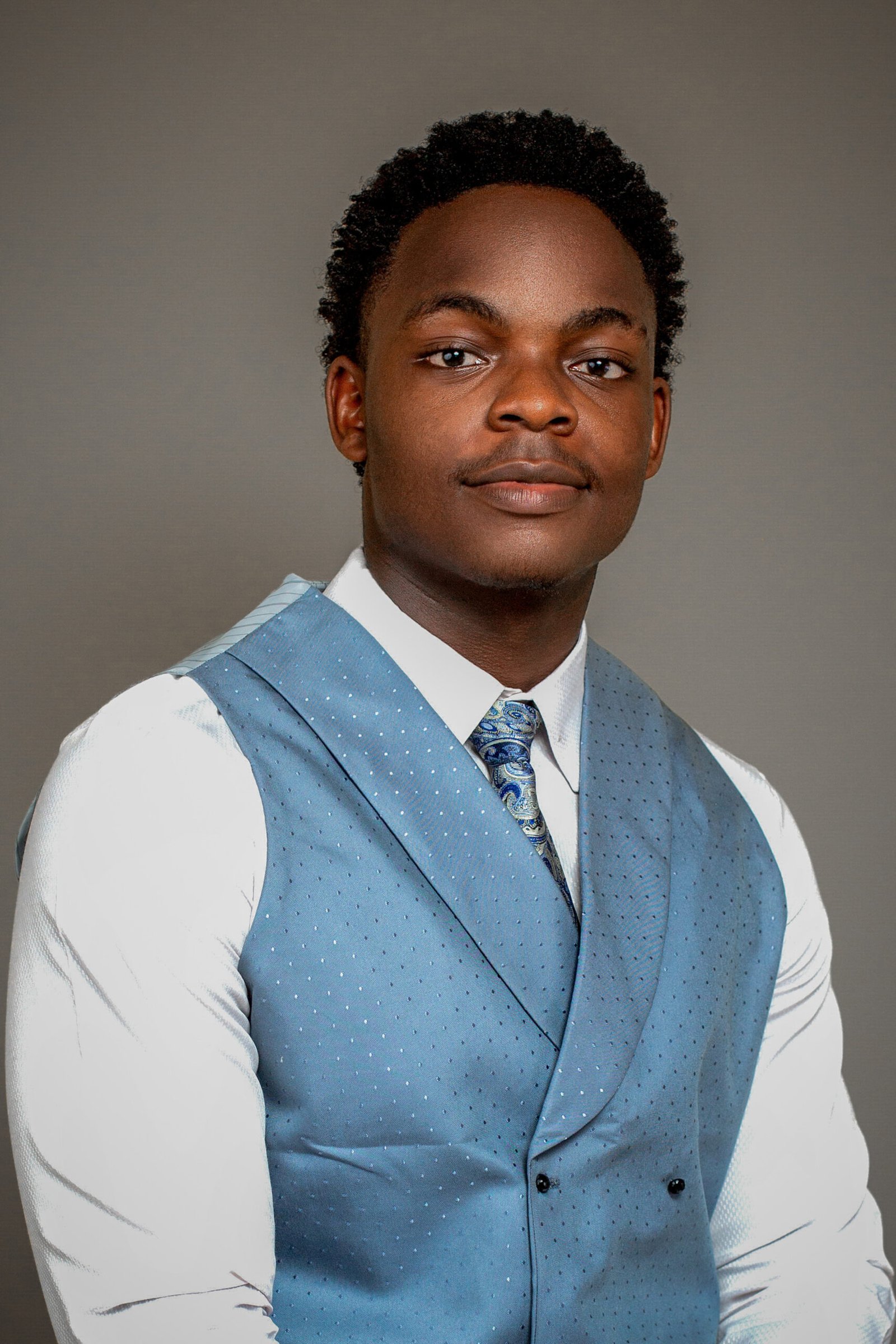 A man wearing a light blue vest, white shirt, and patterned blue tie poses against a plain gray background, looking confidently at the camera with a slight smile.